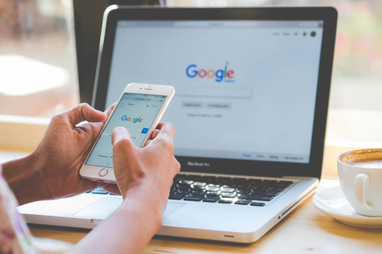 Google on a mobile phone and a mac with coffee next to the comptuer